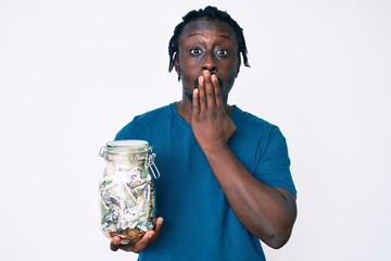 Canvas Print - Young african american man with braids holding jar with savings covering mouth with hand, shocked and afraid for mistake. surprised expression