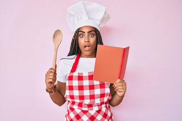 Canvas Print - Young african american woman with braids wearing professional baker apron reading cooking recipe book afraid and shocked with surprise and amazed expression, fear and excited face.