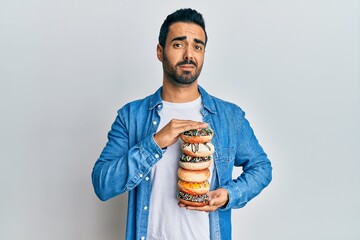 Canvas Print - Young hispanic man holding tasty colorful doughnuts clueless and confused expression. doubt concept.