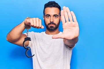 Wall Mural - Young hispanic man holding prisoner handcuffs with open hand doing stop sign with serious and confident expression, defense gesture