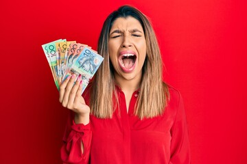 Canvas Print - Beautiful brunette woman holding australian dollars angry and mad screaming frustrated and furious, shouting with anger. rage and aggressive concept.