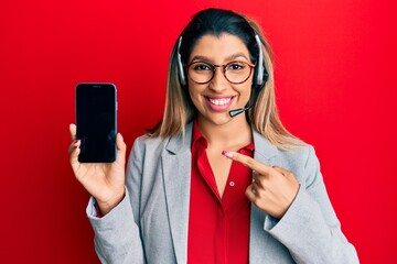 Poster - Beautiful brunette woman wearing operator headset showing smartphone screen smiling happy pointing with hand and finger