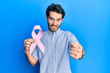 Sticker - Young hispanic man holding pink cancer ribbon annoyed and frustrated shouting with anger, yelling crazy with anger and hand raised