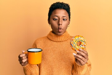 Wall Mural - Young african american girl eating doughnut and drinking coffee making fish face with mouth and squinting eyes, crazy and comical.