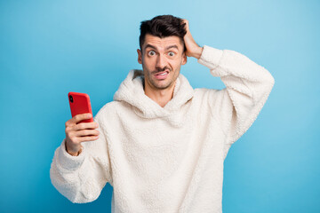 Photo portrait of man scratching head holding phone in one hand wearing wool hoodie isolated on pastel blue colored background