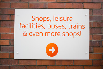 Moderns white sign with orange writing direction customers to shops, leisure facilities, buses and trains with arrow on a brick wall