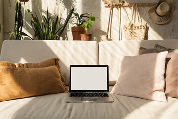 Wall Mural - Laptop with blank screen on comfortable sofa in warm sunlight shadows. Minimal boho styled interior design template with mockup copy space.