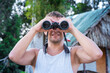 © terezika - Front view of a satisfied young man in a white T-shirt looks through binoculars against the background of a village hut