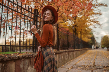 Elegant happy smiling woman wearing stylish orange hat, turtleneck, checkered skirt, holding suede bag with fringe, walking in street of city. Outdoor autumn fashion portrait. Copy, empty space 