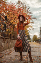 Outdoor Autumn Fashion Full-length Portrait: Elegant Woman Wearing Stylish Orange Hat, Turtleneck, Checkered Midi Skirt, High Boots Holding Suede Bag With Fringe, Walking In Street Of City