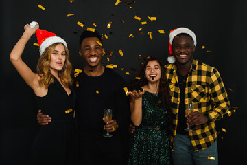 Wall Mural - Four friends of different nationalities throw confetti in santa claus hats and antlers and drink champagne against a black background. A company of young people celebrates Christmas and New Year