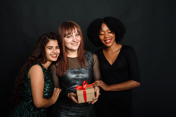 Wall Mural - Three young women of different nationalities are smiling, hugging, holding a present for the new year and christmas on a black background. Gift exchange, giving and receiving concept