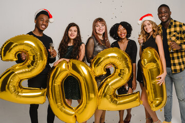 Wall Mural - Cheerful friends are holding numbers 2021 and celebrating New Year by throwing confetti. Cheers. A group of multi-ethnic friends are drinking champagne and having fun on a white background