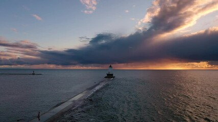 Wall Mural - Beautiful sunset aerial flying over the breakwater pier with the white colored lighthouse sitting at the end in front of the blue and orange colored clouds in the sky beyond as waves splash up.