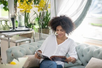 Joyful woman with dark curly hair reading magazine