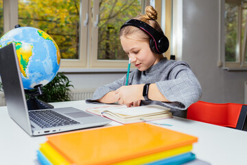 Canvas Print - American teen girl wearing headphones learning language online, using laptop, looking at screen, doing school tasks at home, writing notes, listening to lecture or music, distance education