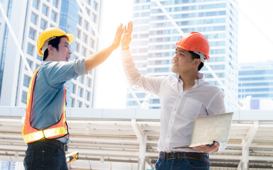 Happy Engineering manager with laptop giving hi five hands with male colleague wear safety jacket and helmet in construction site. Employee or worker greeting, deal,teamwork, collaboration project.