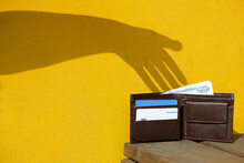 Shadow Of Hand On Yellow Wall Trying To Steal A Leather Wallet With Banknote And Credit Cards. Robber Pickpocket Silhouette. Concept Of Financial Crime, Tax Burden, Unexpected Expenses.