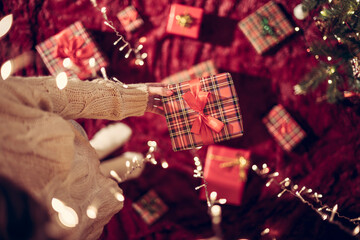 Wall Mural - Woman hand holding out gift box through the glowing garland. Christmas. Top view.