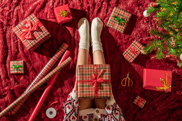 Top view of woman with christmas gift box on knees. Female wrapping gifts for family sitting on fluffy plaid near christmas tree. Flat lay.