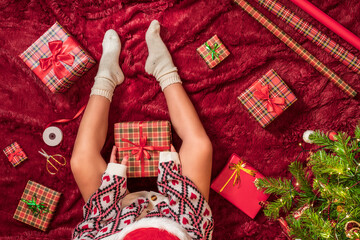 Wall Mural - Top view of woman in red santa claus hat wrapping christmas gifts boxes for family sitting on fluffy plaid near christmas tree. Flat lay.