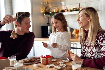 Happy family parents and small child daughter having fun making Christmas cookies decorations at home. Young couple mum and dad helping kid preparing holiday gifts laughing together at kitchen table.