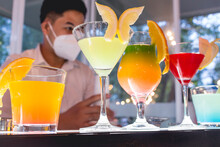 Beautiful Cocktails On Different Colors At A Mobile Bar. A Bartender Wearing A Face Mask Is Behind The Counter. New Normal Situation During An Event.