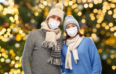 Poster - health, people and winter holidays concept - couple wearing face protective medical mask for protection from virus disease in knitted hats and scarves over christmas tree lights background