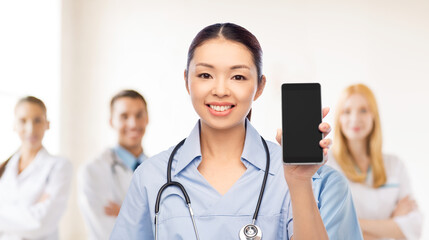 Poster - medicine, profession and healthcare concept - happy smiling asian female doctor or nurse in blue uniform with stethoscope showing smartphone with medical team at hospital on background