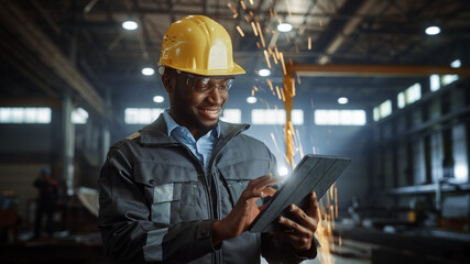 Wall Mural - Professional Heavy Industry Engineer/Worker Wearing Safety Uniform and Hard Hat Uses Tablet Computer. Smiling African American Industrial Specialist Standing in a Metal Construction Manufacture.