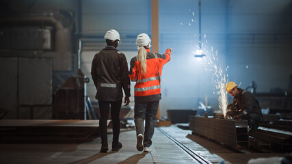 Aufkleber - Two Heavy Industry Engineers Walk Away from Camera in Steel Factory and Discuss Work. Industrial Worker Uses Angle Grinder in the Background. African American Specialist Talks to Female Technician.