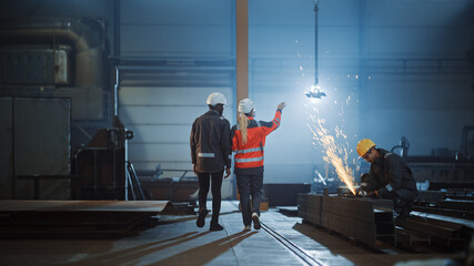 Aufkleber - Two Heavy Industry Engineers Walk Away from Camera in Steel Factory and Discuss Work. Industrial Worker Uses Angle Grinder in the Background. African American Specialist Talks to Female Technician.