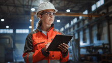 Professional Heavy Industry Engineer/Worker Wearing Safety Uniform And Hard Hat Uses Tablet Computer. Serious Successful Female Industrial Specialist Walking In A Metal Manufacture Warehouse.