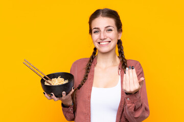 Wall Mural - Young woman over isolated yellow background inviting to come with hand. Happy that you came while holding a bowl of noodles with chopsticks