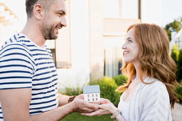 Wall Mural - Side view of happy couple looking at each other while holding statuette of house on blurred background