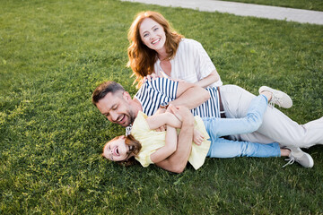 Wall Mural - Happy redhead woman looking at camera near man tickling girl while lying on lawn