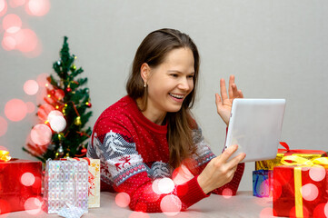 Christmas greetings online. Smiling girl (woman) uses her mobile tablet for communication. Shows the gift to the camera. Video calls to friends, family and parents.