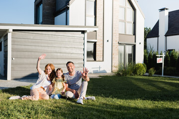 Happy parents with waving hands and daughter sitting on lawn and looking at camera near house