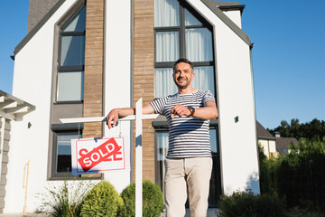 Wall Mural - Happy man with key looking at camera while leaning on sign with sold lettering with modern building on background