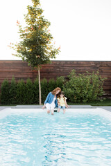 Wall Mural - Cheerful mother and daughter sitting with legs in swimming pool on backyard