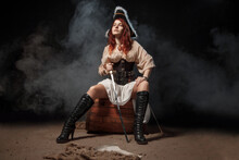 Young Pirate Female With Long Red Hair. Woman Is Wearing A Black Corset Bustier, Tricorn Hat , Gun Belt And Armed With A Pistol And Sword.