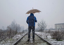 Man Walking On The Railway On A Winter Day. He Has An Umbrella In His Hand. It's Snowing With Big Flakes