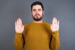 © Roquillo - Serious Young handsome Caucasian man wearing yellow sweater against gray wall pulls palms towards camera, makes stop gesture, asks to control your emotions and not be nervous