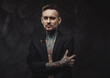 © Fxquadro - Invidivual and stylish guy with tattooed body and modern hairstyle in black suit poses in dark background looking at camera with serious face.