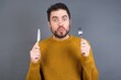 © Roquillo - Model hungry Young handsome Caucasian man wearing yellow sweater against gray wall holding in hand fork knife want tasty yummy pizza pie
