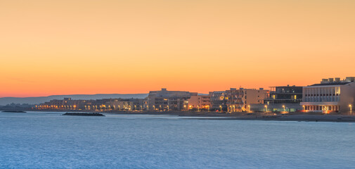 Wall Mural - Vue d'une côte en bord de mer au coucher de soleil avec bâtiments et lumières du soir.