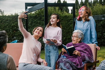 Wall Mural - Hispanic women family reading a book and taking a photo selfie in a backyard outside home in Mexico city