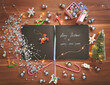 A black paper notebook with Merry Christmas and Happy New Year in silver letters surrounded with colorful Christmas decorations, ornaments and lights on wooden background. Happy holidays background. 