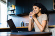 © F8  \ Suport Ukraine - Good looking woman enjoying a cup of coffee while relaxing with her laptop in the kitchen working from home in quarantine lockdown. Social distancing Self Isolation