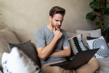 Doubtful young man looking up while working on laptop sit on couch at home